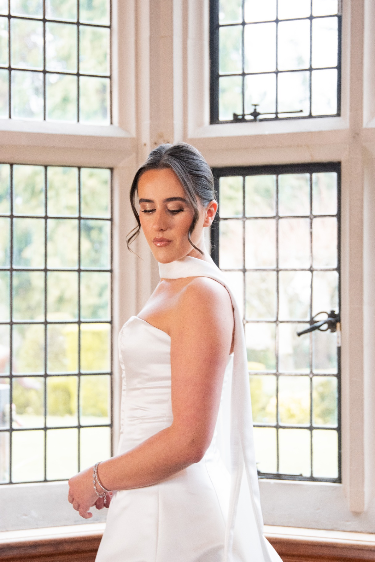 Bride in a satin gown beside a leaded bay window